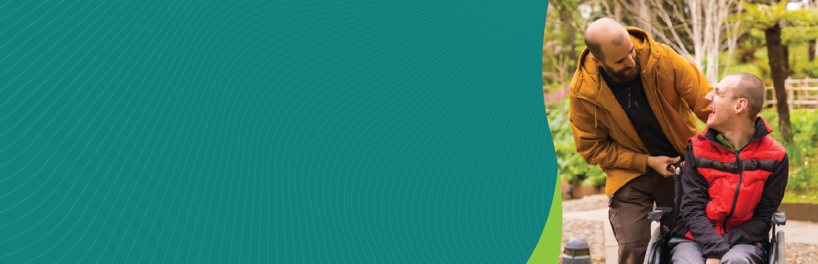 One man smiling at another man in a wheelchair in a park.