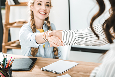 Smiling seated young woman shaking hands with a person standing at her desk