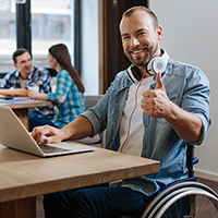 Smiling man in manual wheelchair giving a thumbs up while working on a laptop