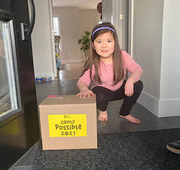 Young girl crouched down beside a box with Camp Possible 2021 sticker on it