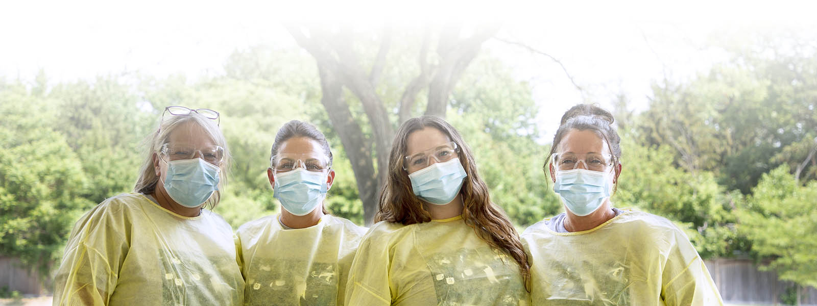 Four smiling MODC staff in protective goggles, blue masks and yellow PPE gowns