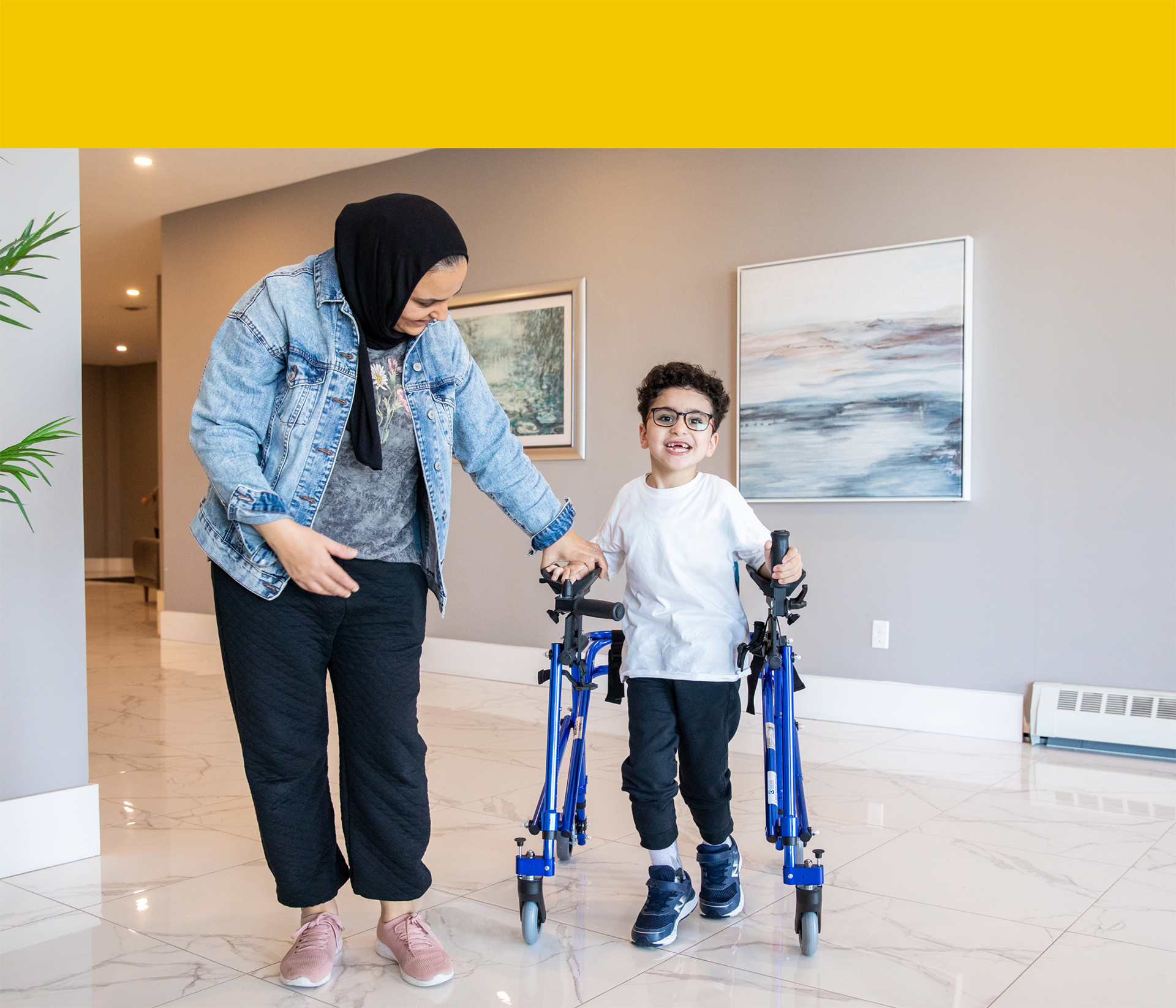Smiling Yousef walking using a supportive walker beside his mother Ola