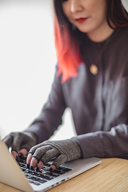 A disabled person types on a laptop while wearing compression gloves. The hands and keyboard are the focal point ©Gregg Jaden
